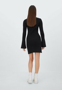Robe midi noire côtelée avec des manches évasées, silhouette ajustée et vue arrière. Le mannequin porte des bottes blanches à semelles épaisses.