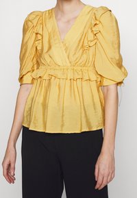 Top giallo, texture con scollatura a V profonda, maniche a volant e vita arricciata. Mostrato abbinato a pantaloni neri.