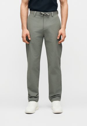 Άνδρας που φοράει γκρι παντελόνι με κορδόνι και λευκά αθλητικά παπούτσια, στέκεται με φόντο ένα απλό φωτεινό σκηνικό.