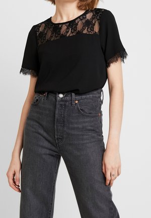 Kobieta ubrana w czarny krótki top z koronkową lamówką na rękawach i dekolcie, zestawiony z wysokim stanem ciemnoszarymi dżinsami.
