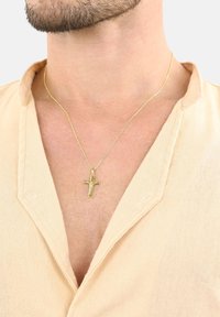 Eine goldene Kreuzkette hängt an einer dünnen Kette und wird über einem hellbeigen Hemd mit Kragen und einem lockeren Ausschnitt getragen.