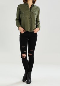 Camisa verde oliva de botones con dos bolsillos frontales, combinada con jeans skinny negros desgastados y botas negras de tobillo. Textura de tela suave.