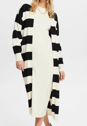 Frau trägt ein langes cremefarbenes Kleid, kombiniert mit einem schwarz-cremefarbenen, breit gestreiften Cardigan und einer goldenen Halskette mit Gliederkette.
