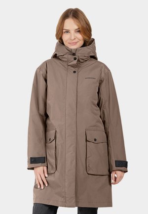 Frau in einem langen taubefarbenen Wintermantel mit Kapuze, Frontklappen-Taschen und schwarzen Handgelenksriemen, lächelt vor einem weißen Hintergrund.