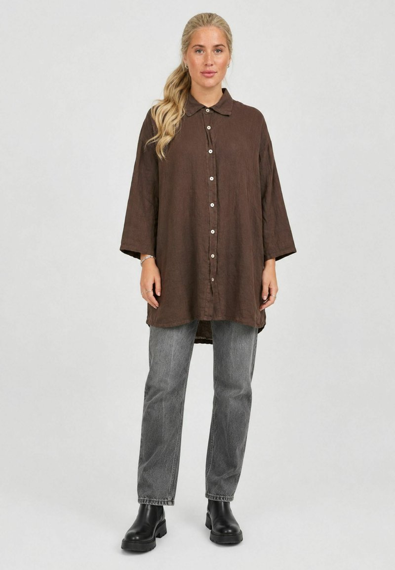 Femme aux cheveux blonds en queue de cheval portant une chemise marron ample à boutons, un jean gris et des bottes noires, debout devant un fond clair uni.