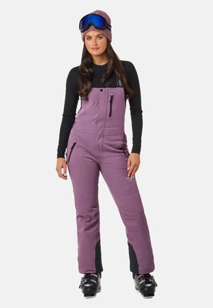 Fialové lyžiarske kombinézy s čiernymi akcentmi, so zipsovými vreckami a nastaviteľnými popruhmi. V kombinácii s čiernym dlhým rukávom a ružovým pleteným čiapkou.