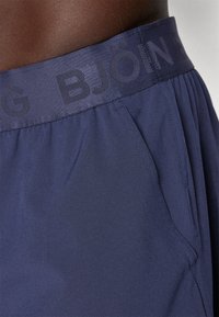 Calções azul-marinho com uma textura suave, apresentando uma cintura elástica larga estampada com "BJÖN." Inclui bolsos laterais e um corte descontraído.