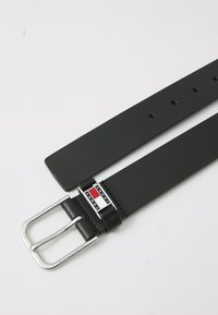 Zwarte leren riem met een zilveren rechthoekige gesp en merklogo met rode, witte en blauwe accenten. Rechte vormgeving met meerdere gaatjes.