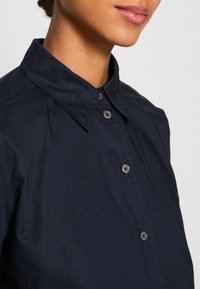 Gros plan d'une personne portant une chemise bleu marine foncé boutonnée avec un col pointu, montrant le cou, le menton et les lèvres.
