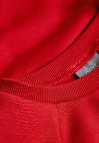 Punane dressipluus soonikkoega kraega, ühtlase tekstuuriga ja sildiga "next". Lähedalt vaadatuna rõhutatakse kangakvaliteeti ja värvi rikkust.