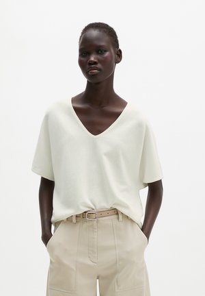 Personne aux cheveux courts portant un haut blanc à col en V et manches courtes, pantalon beige taille haute avec ceinture, mains dans les poches, fond neutre.