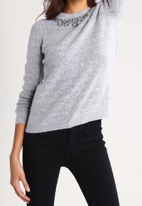 Ljust grå tröja med rundad halsringning och blommiga dekorationer vid kragen. Långärmad och med en något asymmetrisk fåll.