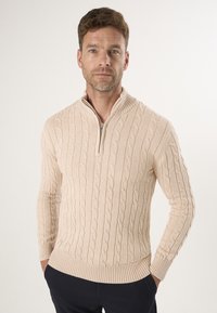 Beżowy sweter dzianinowy z wysokim kołnierzem i półzamkiem, z wzorem w warkocze oraz prążkowanymi mankietami i dołem. Nosi się go na ciemnych spodniach.