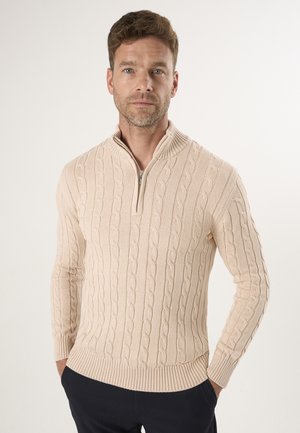 Suéter de punto beige con cuello alto y media cremallera, que presenta un patrón de punto trenzado y puños y dobladillo acanalados. Lleva puesto sobre pantalones oscuros.
