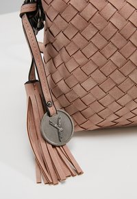 Tkana skórzana torebka w miękkim różowym kolorze z ozdobą w postaci frędzla oraz okrągłą metalową zawieszką z tłoczonym wzorem. Gładka faktura, kompaktowy kształt.