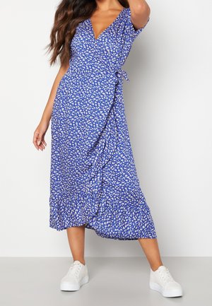 Femme portant une robe portefeuille bleue à imprimé floral blanc avec un ourlet à volants, associée à des baskets blanches à plateforme, debout devant un fond uni.