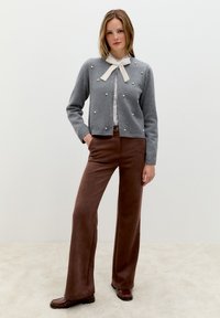 Frau trägt eine graue Strickjacke mit Perlendetails und einer weißen Schleife, ein gestreiftes Shirt, braune weite Hosen und braune Slipper und steht auf einem beigen Teppich.