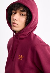 Bordo kapučino gobtuvė pagaminta iš medvilnės mišinio, turi kengūros kišenę ir oranžinį Adidas logotipą. Komplekte yra raišteliai ir erdvus gobtuvas.