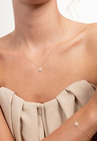 Collier en chaîne d'or avec un pendentif rond orné d'une pierre précieuse, accompagné d'un bracelet assorti. Les deux objets reposent sur une peau nue.