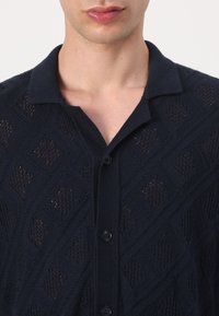 Ung person iført en mørkeblå, tekstureret strikket cardigan med krave og sorte knapper, vist fra nakke til midt bryst.