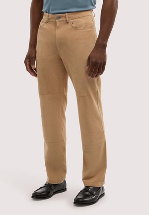 Homme portant un pantalon droit couleur beige et des mocassins en cuir noir, debout devant un fond clair uni.