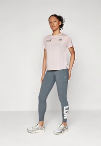 Vaaleanpunainen lyhythihainen urheilupaita, jossa musta logo, yhdistettynä harmaisiin legginsseihin, joissa on sivutasku ja valkoinen teksti. Hopeat väriset urheilukengät.