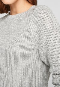 Hellgrauer Strickpullover mit strukturierter Webart, geripptem Halsausschnitt und langen Ärmeln. Nahaufnahme zeigt die Weichheit des Stoffes.