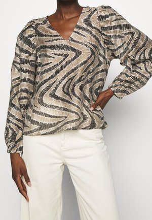 Kvinde iført en langærmet bluse med abstrakt beige og sort zigzag-mønster, kombineret med højttaljede off-white bukser.