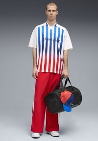 Άνδρας που φορά μπλε, κόκκινη και λευκή ριγέ μπλούζα μοτοσπορτ και κόκκινο παντελόνι, κρατώντας μια μαύρη τσάντα duffel με κόκκινο και μπλε καπέλο.