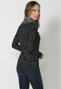 Schwarzes Langarmshirt mit Blättermuster, grauer Stehkragen und taillierte Form, kombiniert mit dunkelblauen Jeans. Texturierte Stoffdetails sichtbar.