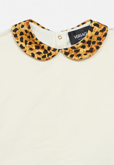 Vêtement blanc avec un col Claudine imprimé léopard jaune et noir et une étiquette Versace à la fermeture par bouton au niveau du col.