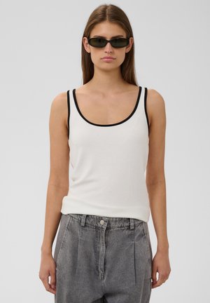 Femme portant des lunettes de soleil noires, un débardeur blanc avec une bordure noire et un jean gris taille haute, debout devant un fond uni.