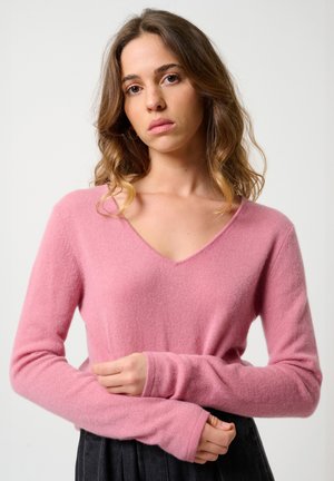 Jeune femme aux cheveux ondulés portant un pull rose en V et un pantalon sombre, se tenant les bras croisés sur un fond uni.