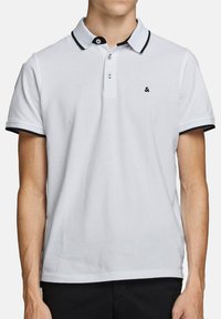 Polo blanc en coton, avec un col noir et des finitions de manches noires, une patte de boutonnage à deux boutons, et un petit logo sur la poitrine.