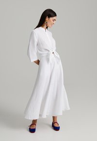 Bela lanena obleka s tremi četrtinskimi rokavi, zavezana v pasu. Model nosi pisane viseče uhane in modre ter rdeče ravne čevlje.