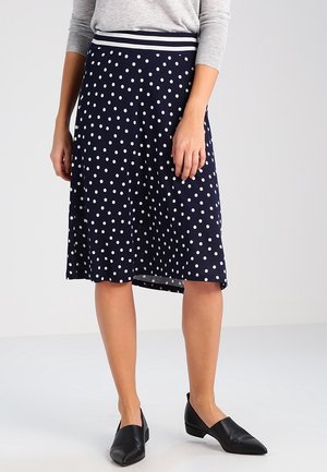 Kobieta ubrana w granatową spódnicę do kolan w białe kropki, szary top i czarne spiczaste buty, stojąca na tle jednolitego tła.