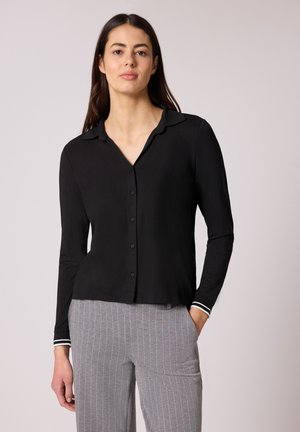 Femme portant une chemise noire longue à manches longues boutonnée et un pantalon gris à fines rayures, une main dans la poche, sur un fond uni.