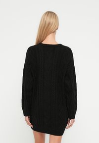Zwarte gebreide jurksweater met getextureerde kabelpatronen, lange mouwen en een geribbelde zoom. Achteraanzicht met een brede halslijn.