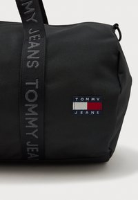 Czarna torba sportowa wykonana z wytrzymałego materiału, z wyraźną naszywką z logo oraz teksturowanym paskiem z literami "TOMMY JEANS". Zapięcie na zamek.