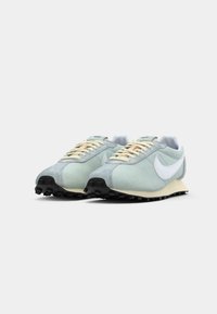 Par nogavic z jasno modro barvo Nike s belimi logotipi swoosh, smetanastimi vezalkami in črnimi gumijastimi podplati na enobarvni beli podlagi.