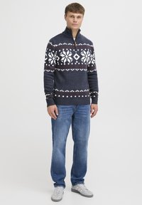 Gebreide trui in marineblauw met een wit en bordeaux sneeuwvlokpatroon, kwart-rits kraag, geribbelde manchetten en zoom, gecombineerd met blauwe jeans en grijze schoenen.