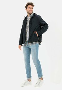 Dark blue hooded jacket with a zippered pocket, over a light plaid shirt, paired with light blue jeans and white sneakers.
