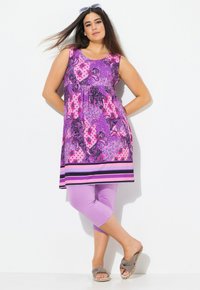 Fialové a růžové vzorované bezramínkové tunikové šaty se staženým pasem, kombinované s levandulovými capri legínami a neutrálními sandály.