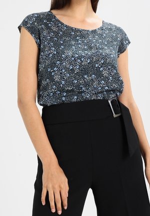Femme portant un chemisier noir à manches courtes avec un motif floral, rentré dans un pantalon noir taille haute avec une large ceinture et une boucle rectangulaire.