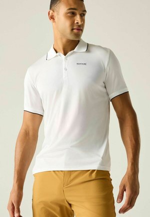 Mann trägt weißes Poloshirt mit kurzen Ärmeln und schwarzer Einfassung sowie Khakihose, steht vor einfarbig hellem Hintergrund.