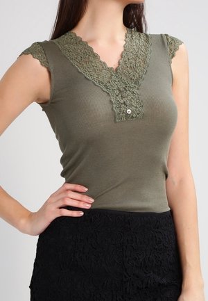 Frau trägt ein ärmelloses, olivgrünes Oberteil mit Spitzen-V-Ausschnitt und Knopfdetails, kombiniert mit einem schwarzen Spitzenrock, Hand in der Hüfte.