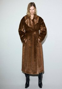 Cappotto lungo in pelliccia sintetica marrone con ampio colletto, chiusura a doppi petti, cintura e tasche frontali. Indossato sopra stivali neri alti fino al ginocchio.