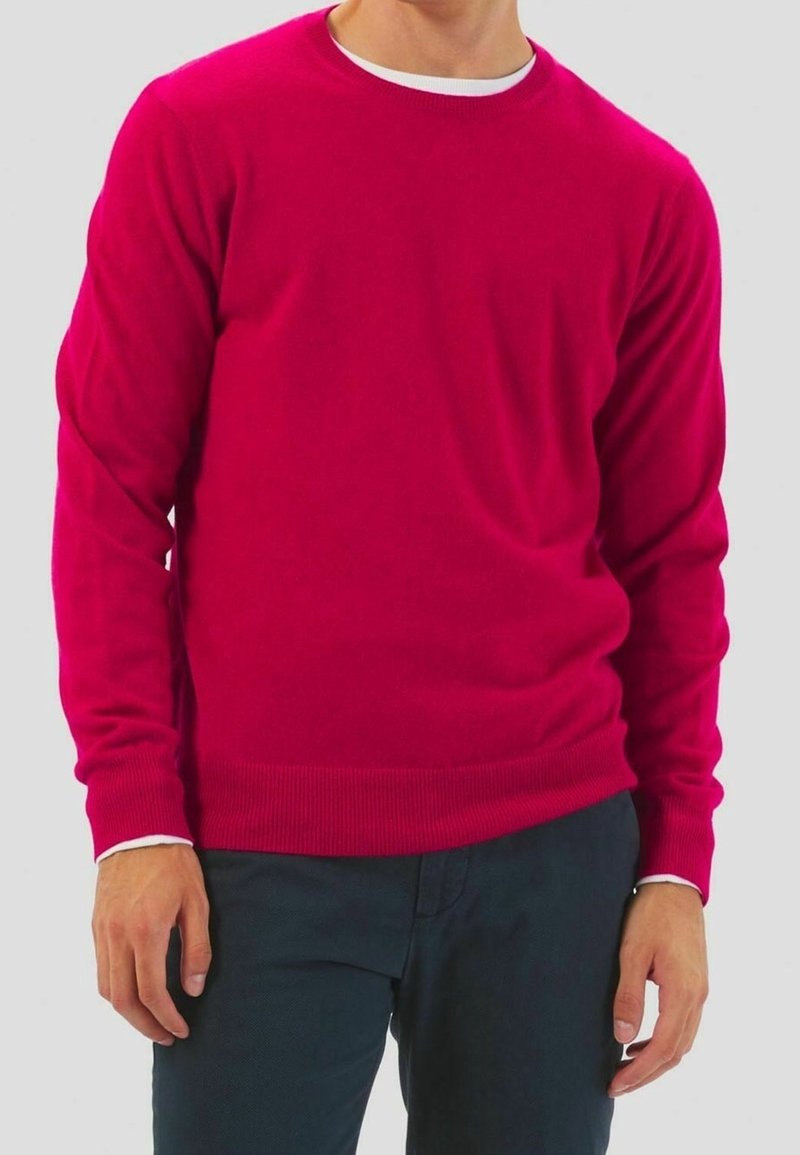 Homme portant un pull rouge vif à manches longues avec un maillot blanc et un pantalon foncé, debout devant un fond clair.