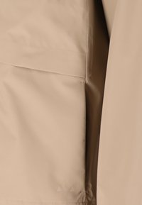 Beige stof med synlige sømme og syninger, muligvis en del af et beklædningsgenstand eller polstring med glat tekstur og lette folder.