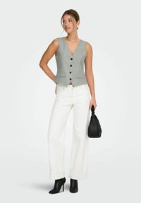Femme portant un gilet sans manches gris à boutons, un pantalon blanc à jambes larges, des bottes à talons noires, tenant un sac à main noir sur un fond blanc.